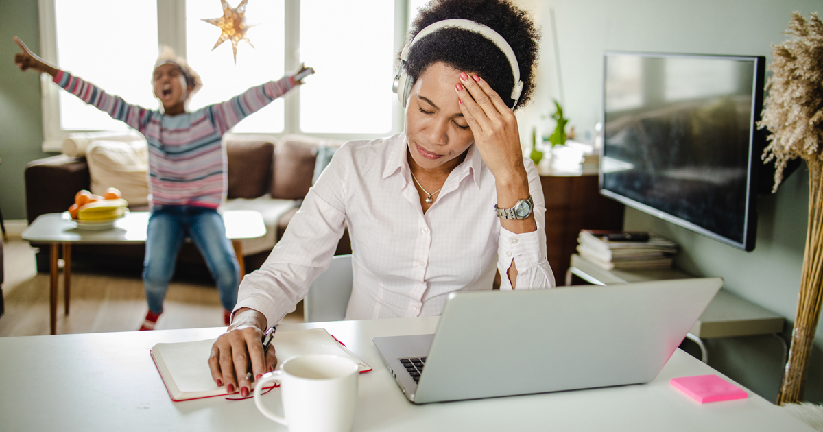 Woman at computer looking stressed. Child playing in background.