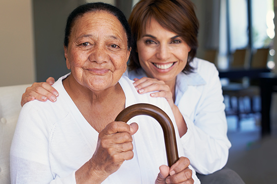 A woman sitting on the couch holding her cane. Her daughter is behind her, with her hands on her shoulders. 