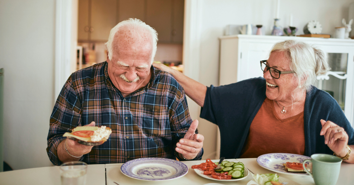 An older couple eating breakfast and laughing at the kitchen table