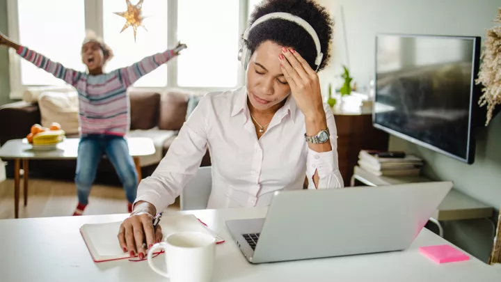 Woman at computer looking stressed. Child playing in background.