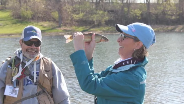 Woman next to a lake holding a fish up to her face and smiling while a smiling man watches