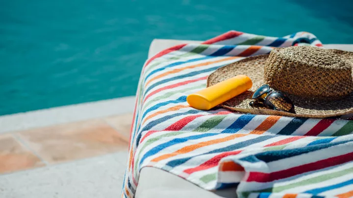 A sun hat, sunglasses, and a bottle of sunscreen on top of a towel on a chair by a pool