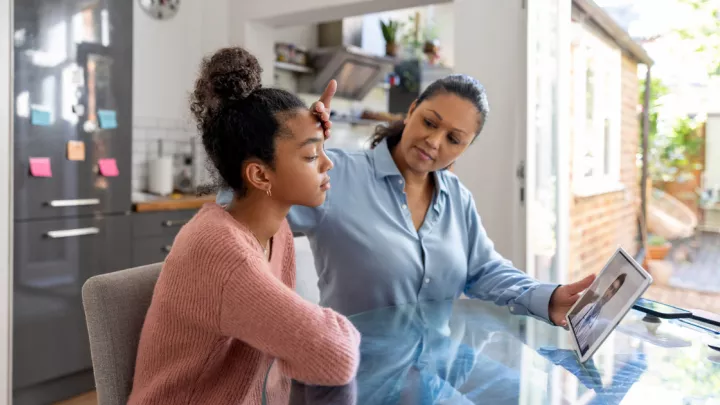 Mother feeling her teen daughter's forehead while on a telehealth appointment with a doctor on a tablet